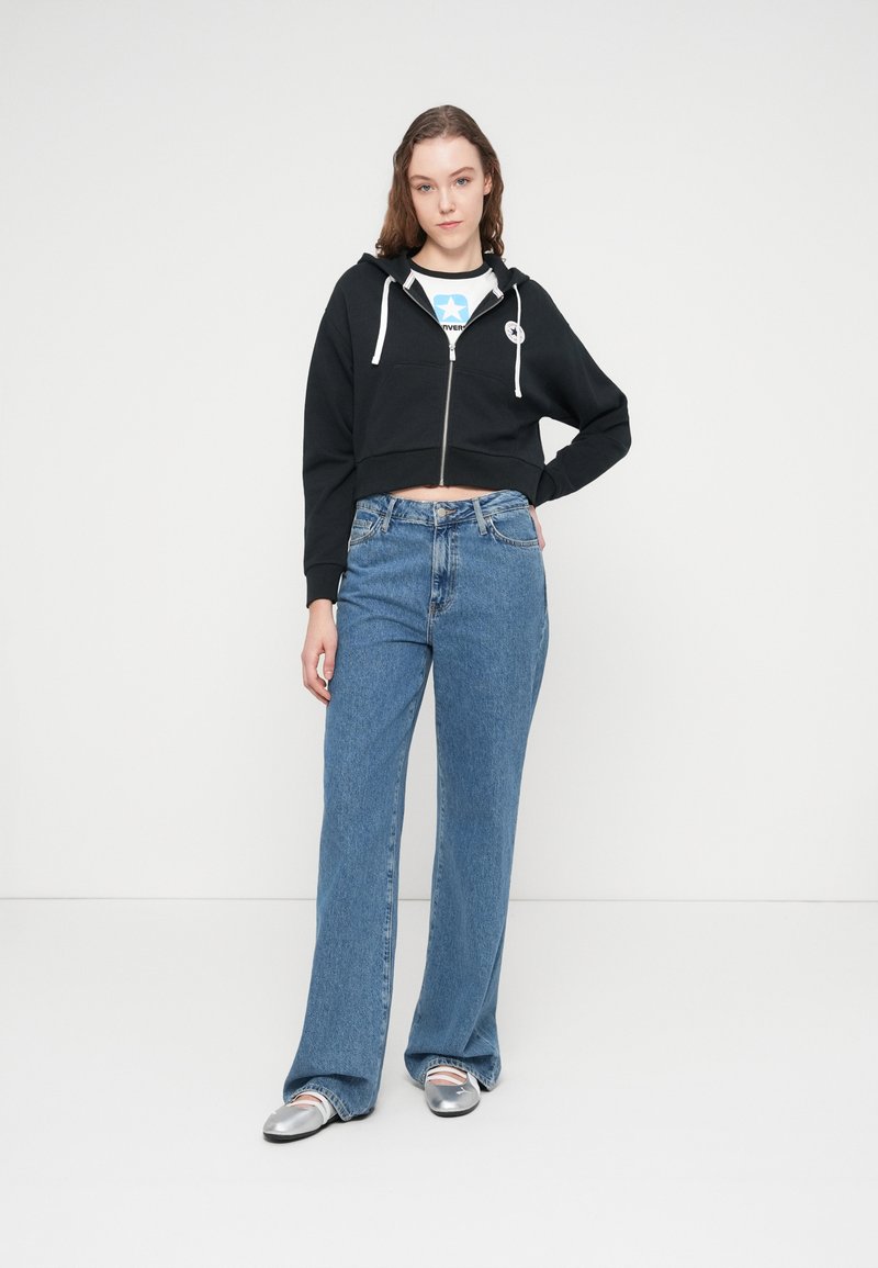 Sort cropped hættejakke med lynlås og hvide snore, kombineret med højttaljede blå jeans. Modellen har sølvfarvede flade sko på. Enkel, afslappet outfit.