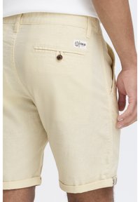 Khaki shorts met opgerolde omslagen, voorzien van een knoopzak aan de achterkant en een label op de tailleband. De stof heeft een gestructureerde uitstraling.