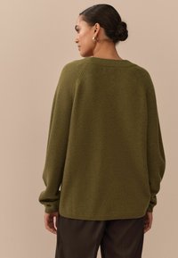 Olive green oversized sweater with ribbed neckline and cuffs, featuring a smooth texture and seam detailing, worn over dark trousers.