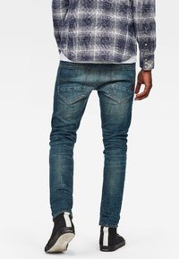 Jeans de denim azul ajustados con áreas descoloridas, bolsillos traseros y acentos de costura, combinados con zapatos negros con rayas blancas.