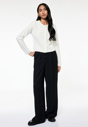 Kobieta z długimi czarnymi włosami, ubrana w biały kardigan na guziki, czarne spodnie o szerokich nogawkach i czarne buty, stojąca z jedną ręką na biodrze.