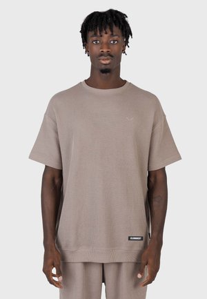 Mand med korte dreadlocks iført løs beige tekstureret kortærmet skjorte og matchende bukser, stående foran en ensartet lys grå baggrund.