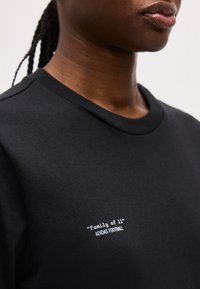 Κοντινό πλάνο ατόμου που φοράει μαύρο μπλουζάκι Adidas με λευκά γράμματα που γράφουν «Οικογένεια των 11» Adidas Football στο στήθος.