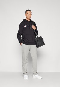 Felpa nera con cappuccio e logo bianco, pantaloni della tuta grigi e sneakers bianche. Borsa nera con tracolla. Outfit semplice e casual da atletica.