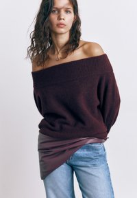 Ung kvinde med bølget hår iført en off-shoulder mørk lilla sweater, asymmetrisk lædernederdel og lyseblå jeans mod en ensfarvet baggrund.