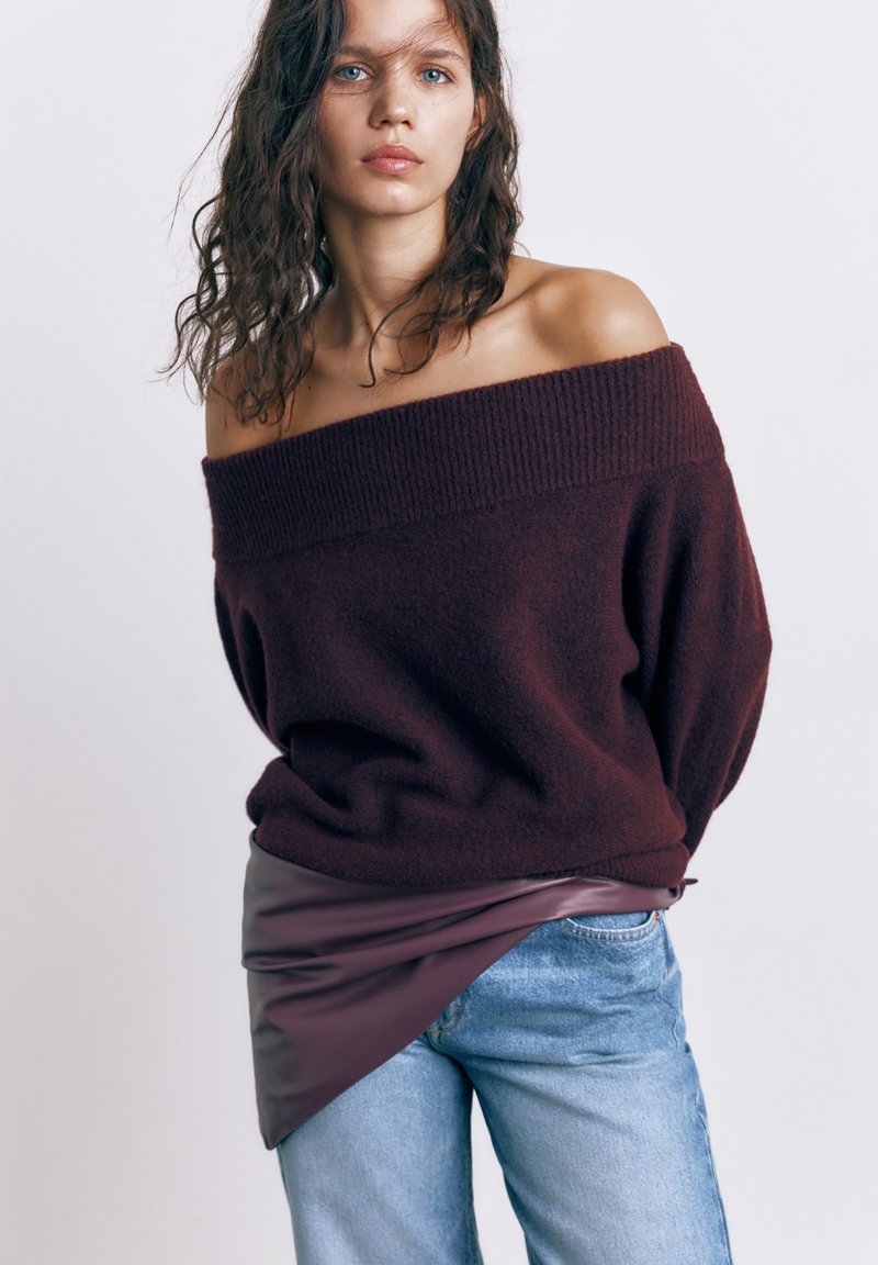Ung kvinde med bølget hår iført en off-shoulder mørk lilla sweater, asymmetrisk lædernederdel og lyseblå jeans mod en ensfarvet baggrund.