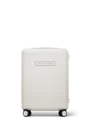 Valise rigide de couleur blanche mate avec des stries horizontales, une poignée supérieure et quatre roues noires pour la mobilité. L'avant présente une ouverture étroite.