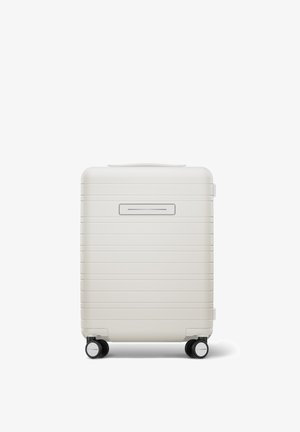 Valise rigide de couleur blanche mate avec des stries horizontales, une poignée supérieure et quatre roues noires pour la mobilité. L'avant présente une ouverture étroite.