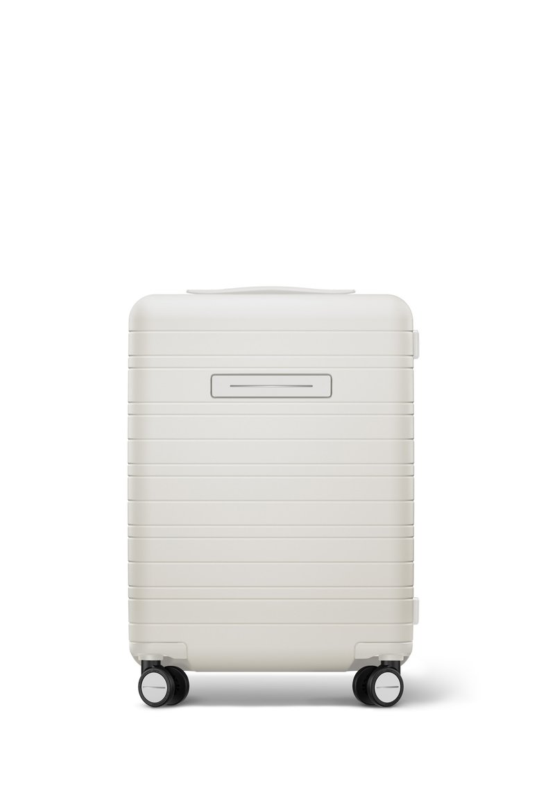 Hartschalenkoffer in mattem Weiß mit horizontalen Rillen, Tragegriff und vier schwarzen Rollen für Mobilität. An der Vorderseite befindet sich eine schmale Öffnung.