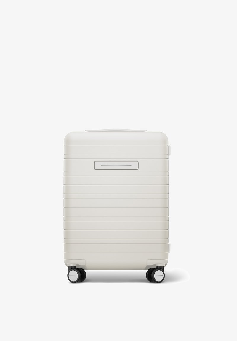 Hartschalenkoffer in mattem Weiß mit horizontalen Rillen, Tragegriff und vier schwarzen Rollen für Mobilität. An der Vorderseite befindet sich eine schmale Öffnung.
