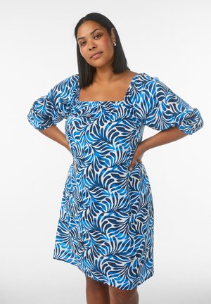 Žena s rovnými čiernymi vlasmi má na sebe modrobiele vzorované šaty po kolená s naberanými rukávmi a stojí s rukami v bok.