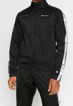 Mężczyzna ubrany w czarny dres Champion z białymi paskami z logo na rękawach i spodniach, stojący na jednolitym tle.