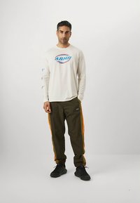 Kremasta košulja s dugim rukavima s printom logotipa; maslinasto zelene hlače s narančastim bočnim prugama; crne tenisice; stoji na jednobojnoj pozadini.