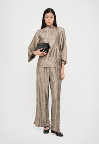 Haut beige texturé avec des manches larges et un pantalon ample assorti ; le mannequin tient une pochette noire ; chaussures noires avec des accents.