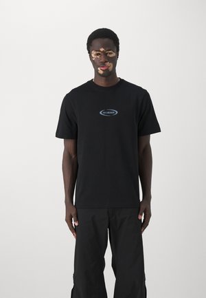Czarny krótki t-shirt z bawełny z niebieskim logotypem na piersi. Model nosi luźne czarne spodnie cargo.