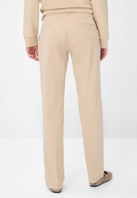 Beige lige ben bukser med en glat tekstur, der har to baglommer og en subtil plade langs forsiden, parret med lyse sko.