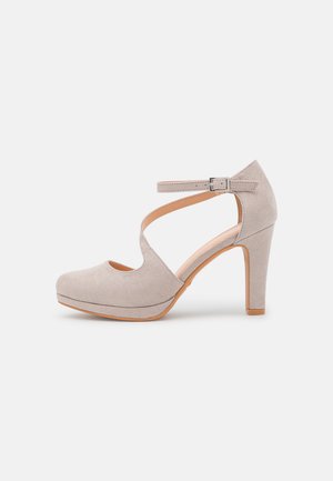 Beige suede high heel shoe with an ankle strap, rounded toe, platform front, and cut-out design on upper. Wood-like heel, smooth texture.