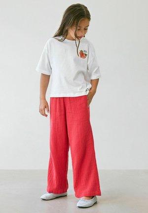 Młoda dziewczyna nosząca białą koszulkę z nadrukiem truskawki i luźne czerwone spodnie, patrzy w dół z ręką w kieszeni na jednolitym tle.