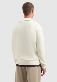 Mann mit kurzem Haar, der einen gerippten, cremefarbenen Pullover, beiger Hose und dunkelunterem T-Shirt trägt, von hinten vor einem grauen Hintergrund abgebildet.