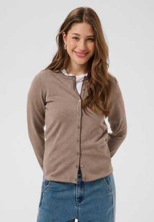 Joven mujer con pelo largo y castaño que lleva un cárdigan beige abotonado sobre una camiseta blanca y vaqueros azules, sonriendo con las manos detrás de la espalda.