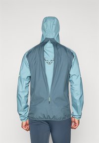 Jachetă windbreaker cu glugă, având un design bicolor în nuanțe de albastru deschis și închis, material texturat și un logo pe spate.