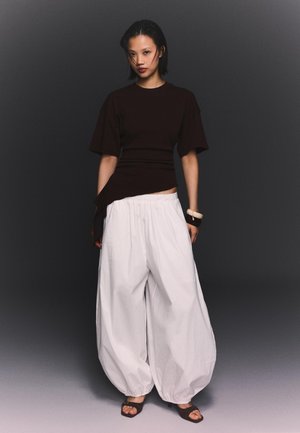 Vrouw staat tegen een donkere achtergrond, draagt een zwart shirt met korte mouwen, losse witte broek, donkere sandalen en armbanden aan haar rechterpols.