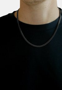 Metal kæderekke med et tykt led design, der har en sort, blank finish, båret over en simpel sort t-shirt.