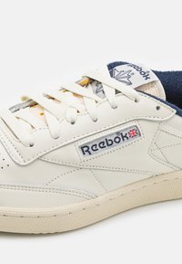 Klassiske Reebok-sneakers i off-white lær med blå detaljer, teksturert gummisåle og synlig logo; har flate lisser og perforert tåboks.