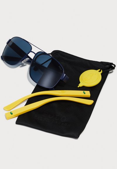Ochelari de soare navy cu lentile închise, însoțiți de două vârfuri galbene ale tâmplelor și o etichetă galbenă, prezentati într-un sac negru cu șnur.