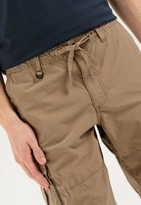Close-up of a person wearing beige cargo shorts with a tied waist drawstring and a dark shirt, hand resting on the shorts pocket.