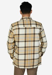 Mann von hinten gesehen, trägt ein beige-weiß-braunes kariertes Hemd mit langen Ärmeln und braune Hose vor weißem, einfarbigem Hintergrund.