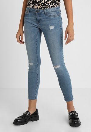 Femme portant un jean skinny déchiré bleu clair, des mocassins noirs avec des chaînes argentées, et un haut à motif léopard visible à la taille.
