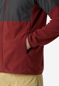 Persona vistiendo una chaqueta de forro polar en dos tonos, rojo y gris oscuro, con la mano en el bolsillo lateral con cremallera, combinada con pantalones de un beige claro.