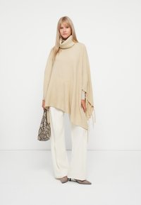 Poncho de malha bege com colarinho alto, textura canelada e acabamentos com franjas, combinado com calças largas brancas e uma bolsa de estampado de cobra.