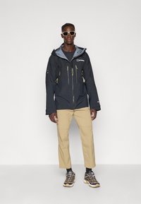 Black waterproof jacket with gray lining, zippered pockets with yellow accents. Paired with beige pants and black hiking shoes.