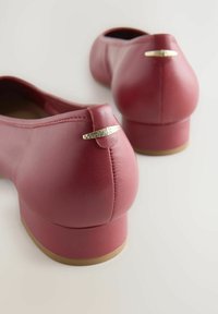 Zapatos planos de cuero rojo con punta redonda y tacón bajo. Presentan un acento dorado cerca del tacón. Textura suave con costuras mínimas.