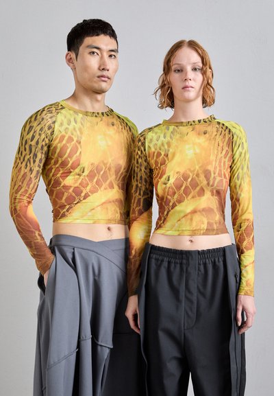 Camisetas cortas de manga larga en estampado de serpiente amarillo y marrón. Combinándolas con pantalones grises drapeados que presentan cinturillas anchas y bolsillos.