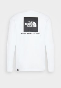 Vit långärmad skjorta med The North Face-logotyp och texten "Never Stop Exploring" tryckt i svart på baksidan.