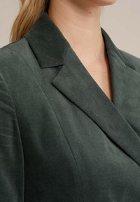 Sacou verde închis cu guler cu revere și textură moale. Vedere de apropriere care evidențiază detaliile materialului și ale reverului structurat.