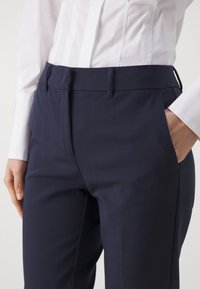 Pantalon sur mesure bleu marine avec une texture lisse, devant plat, passants pour ceinture et poches latérales. Assorti à une chemise blanche à manches longues.