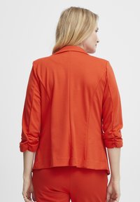 Oranžový blazer s dobře střiženým designem, měkkou texturou a ohrnutými rukávy. Má klopový límec a jemné švy na zadní straně.