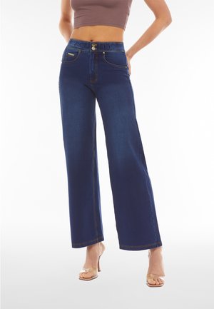 Jeans a gamba larga di colore blu scuro con vita alta, due tasche frontali, cuciture dorate e due chiusure a bottoni frontali. Sandali con tacco traslucidi.