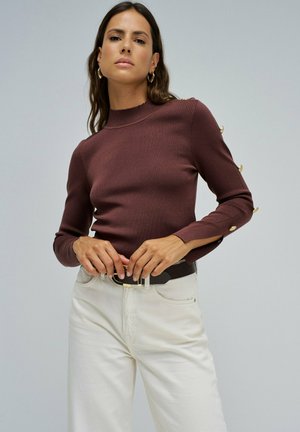 Top de manga larga burdeos con cuello alto y detalles de botones en las mangas, combinado con pantalones blancos de talle alto y un cinturón negro.