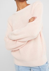 Pull tricoté rose clair à coupe ample, large encolure bateau et poignets côtelés. Le design texturé ajoute un intérêt visuel.