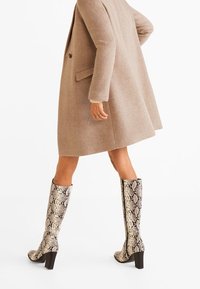 Personne portant un manteau beige et des bottes à talons hauteur genou avec un motif en peau de serpent marchant sur un fond blanc.