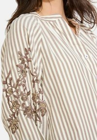 Blusa a righe beige e crema, con ricamo floreale sulla manica arricchita da paillettes. Collo con bottoni e vestibilità comoda.