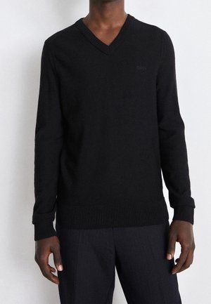 Mężczyzna w czarnym swetrze z dekoltem w serek i ciemnych spodniach, stojący na tle jasnego, jednolitego tła.