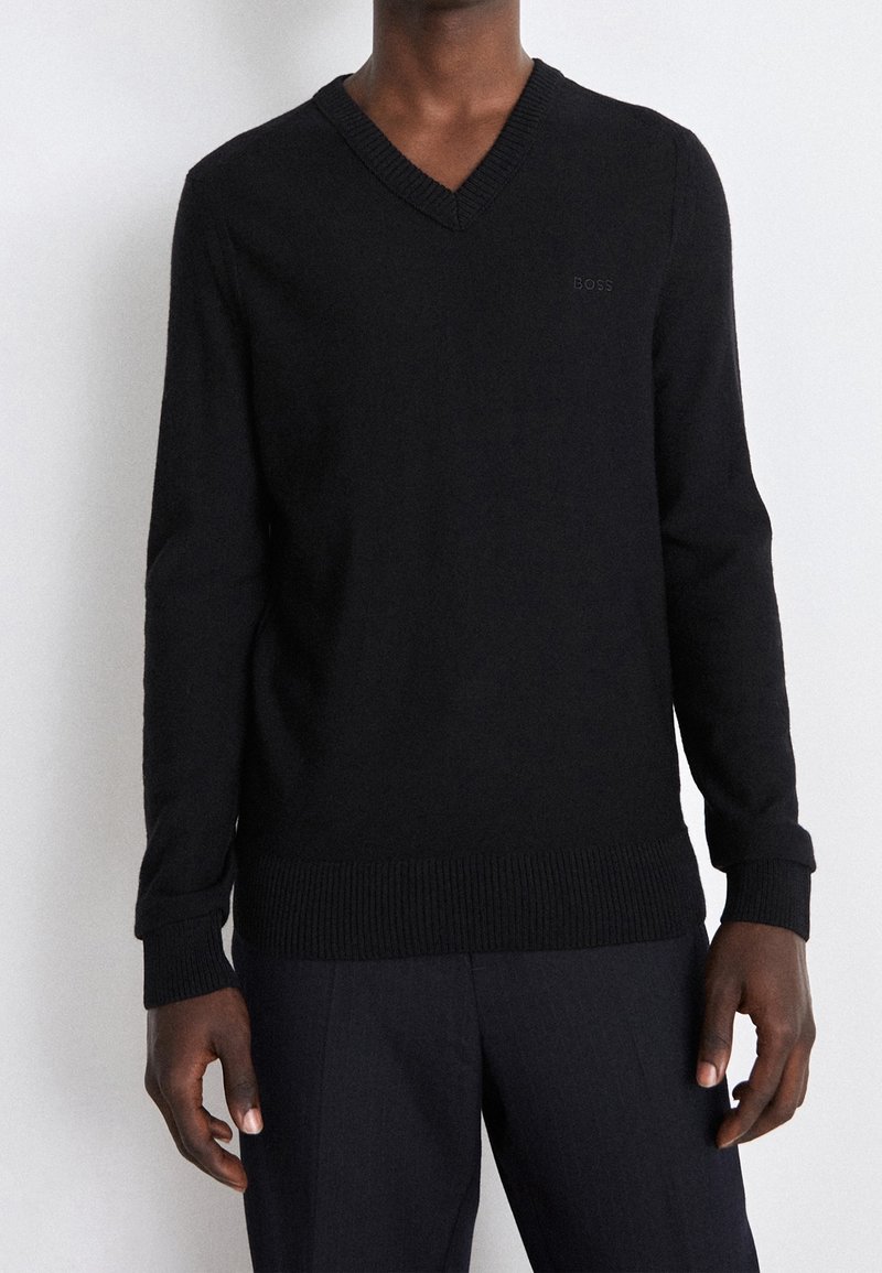 Homme portant un pull noir à encolure en V et un pantalon sombre, debout devant un fond clair uni.