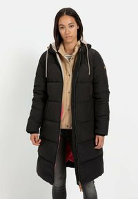 Lange zwarte pufferjas met capuchon, rits aan de voorkant en trekkoorden. Gevoerd met roze stof, gelaagd over een beige fleece sweater en gestreept shirt.