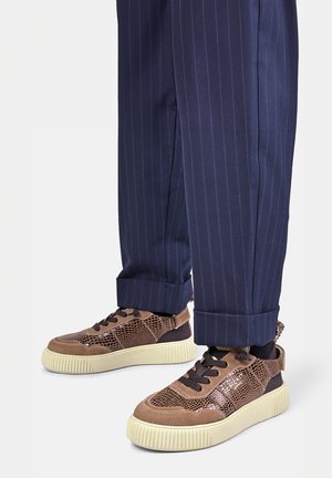 Bruine gestructureerde sneakers met zwarte veters, gedragen met marineblauwe pinstripe-opgerolde broek, tegen een eenvoudige achtergrond.